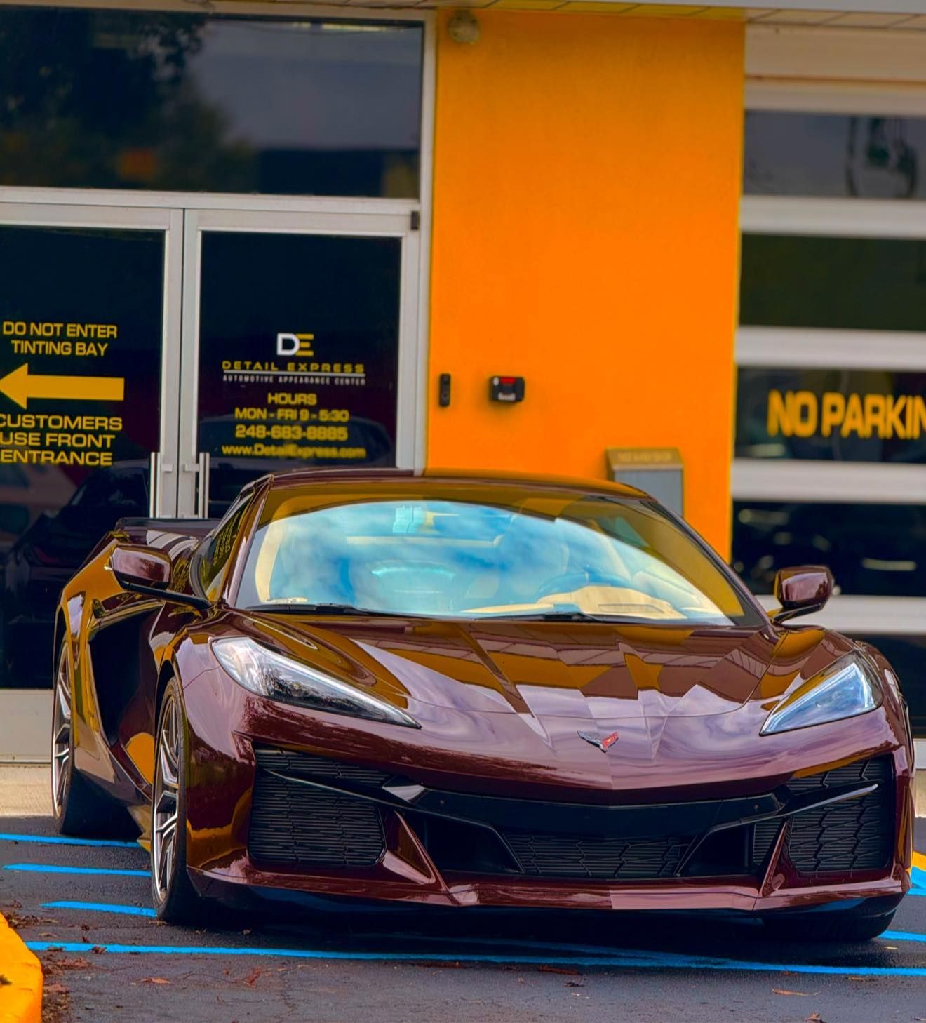 Dark maroon sports car parked in front of an orange storefront with a “No Parking” sign.