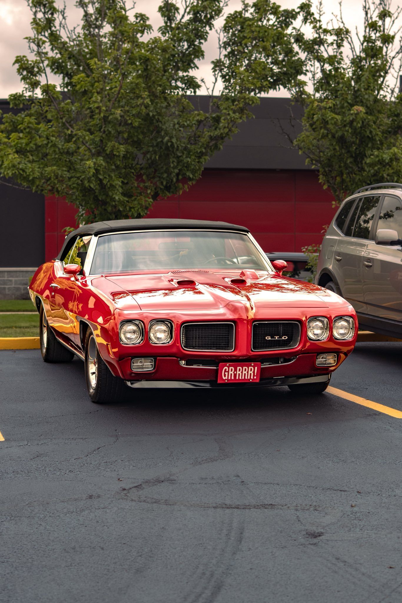 Red classic muscle car parked in a lot, front view, with trees and a red garage behind it
