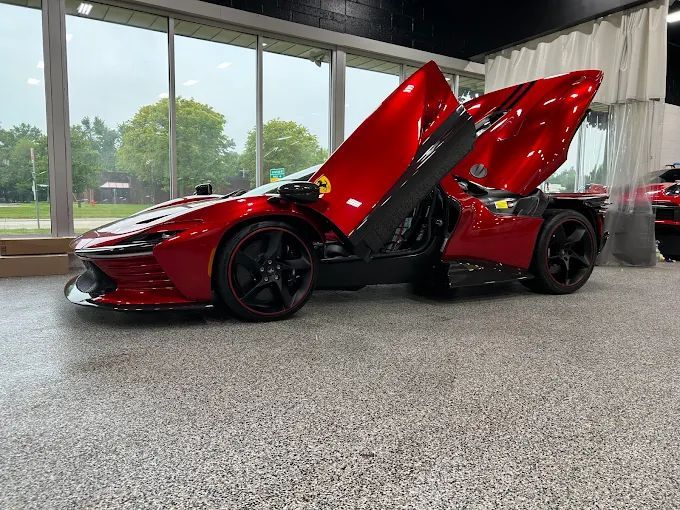 Red sports car with gullwing doors open in a showroom, on a gray floor with curtains and trees outside.