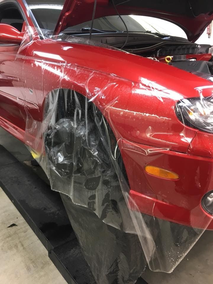 Red car in a shop with front left wheel and fender covered in clear plastic protection film.