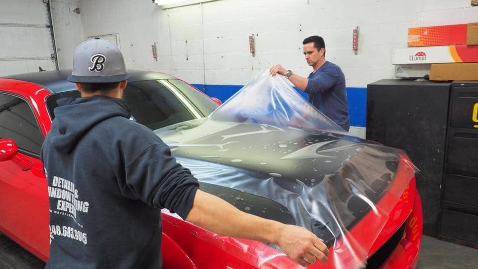 Two people wrapping a red sports car in clear protective film inside an auto shop
