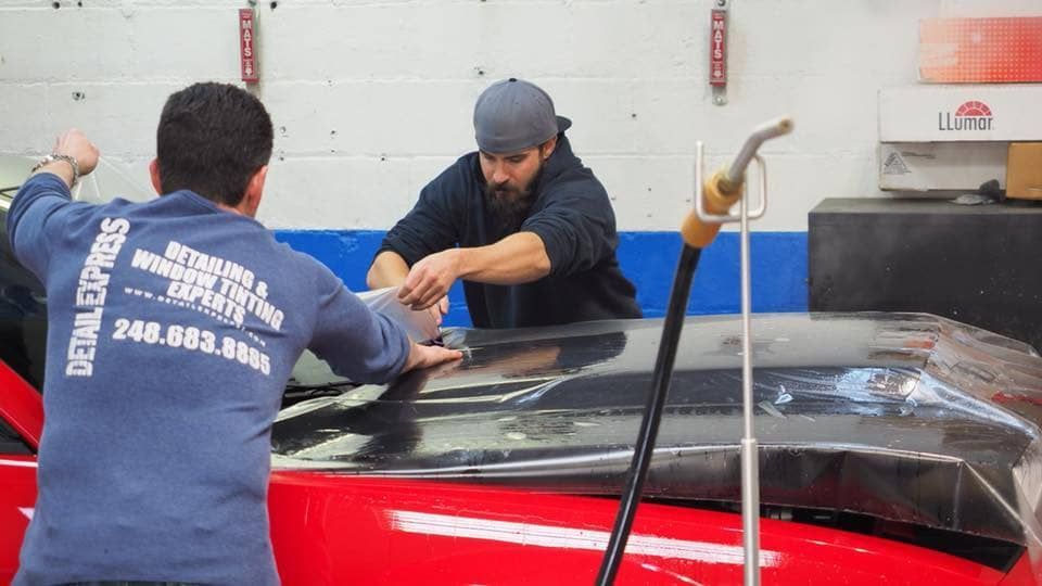 Two workers in a shop handling a large black and red car part with tools on a workbench