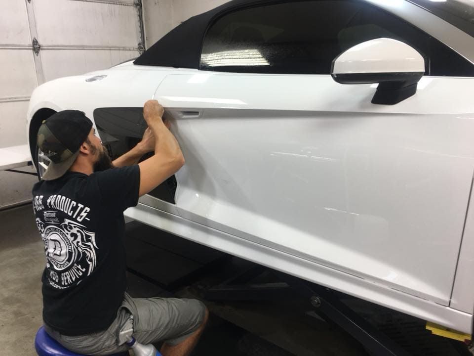 Person kneeling beside a white sports car, applying film or detailing the side panel in a garage