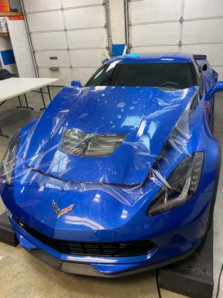 Blue sports car in a garage, front hood damaged and covered with a clear plastic sheet.