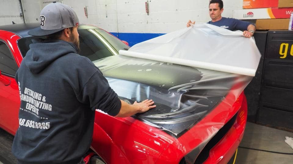 Two workers wrap a red car in clear protective film inside a garage.
