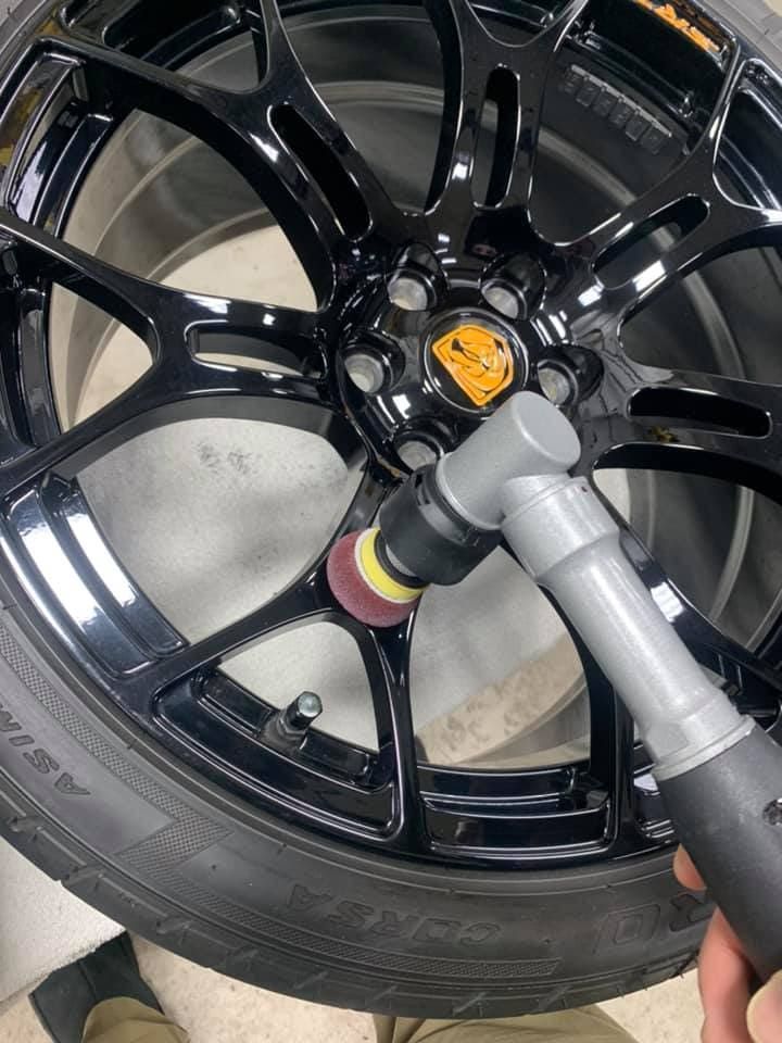 Close-up of a glossy black car wheel being polished with a handheld buffer.