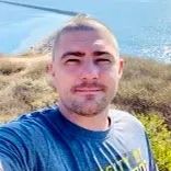 Person taking a selfie outdoors by the water, with dry grass and blue sky in the background.