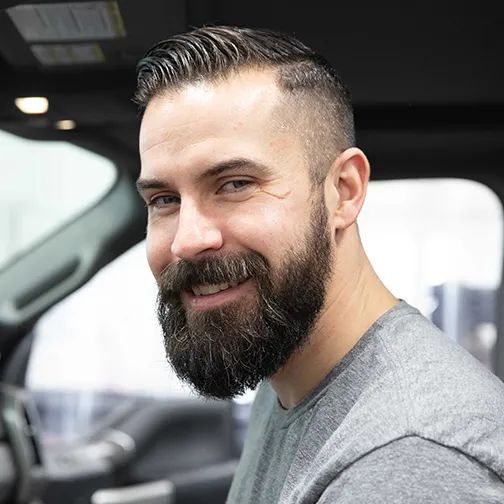 Smiling man with a beard sitting in a car, wearing a gray shirt.
