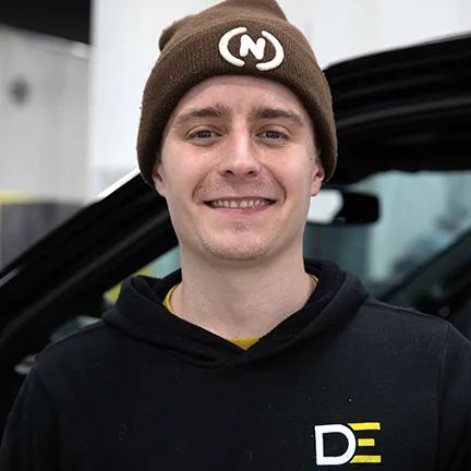 Smiling person in a brown beanie and black hoodie standing beside a vehicle.