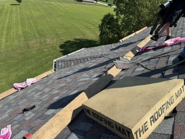 Roofing work on a sloped shingle roof, with workers and roofing materials beside a grassy yard.