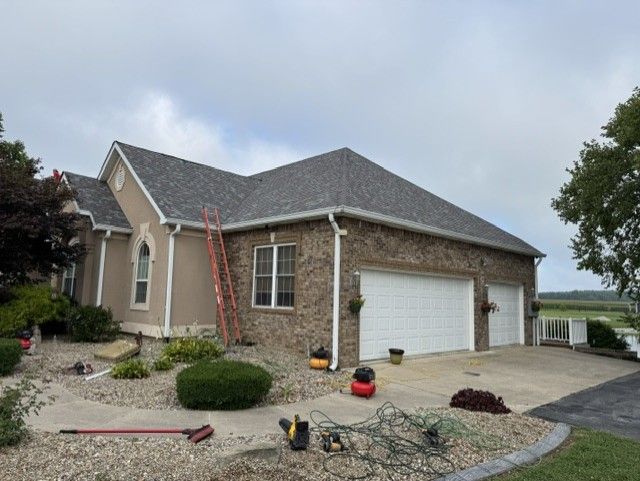 Brick suburban house with white garage, ladder, and yard tools on a driveway under cloudy sky