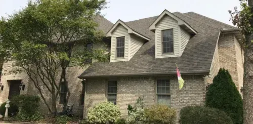 A large brick house with a gray roof and white trim is surrounded by trees and bushes.