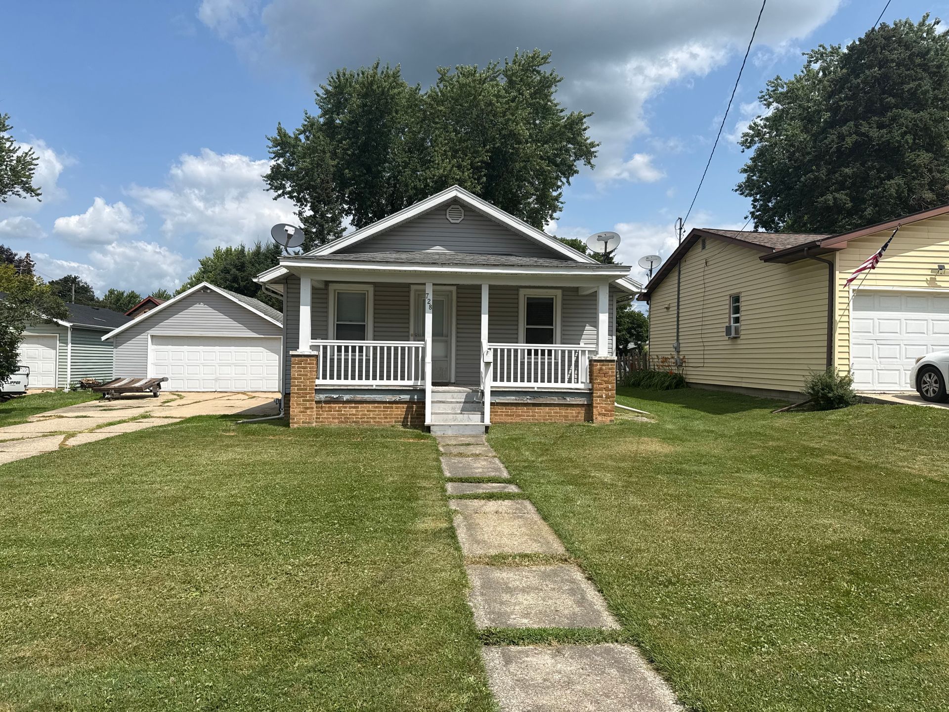 Small white house with front porch and brick base, centered behind a concrete walkway under a blue sky.