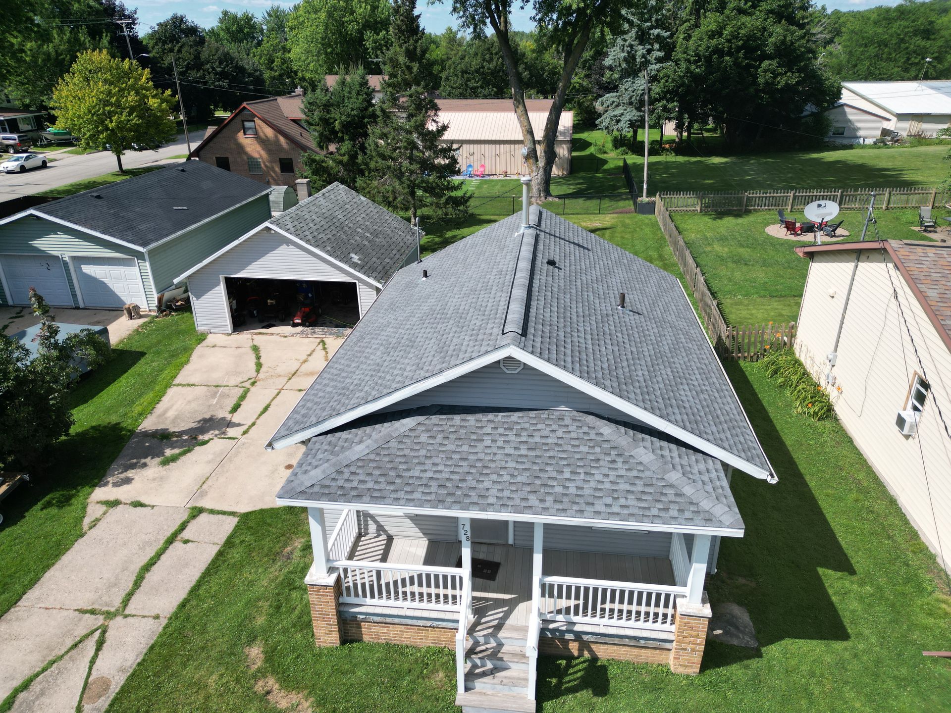 Aerial view of a gray-roofed house with a front porch, driveway, and garage in a suburban neighborhood.