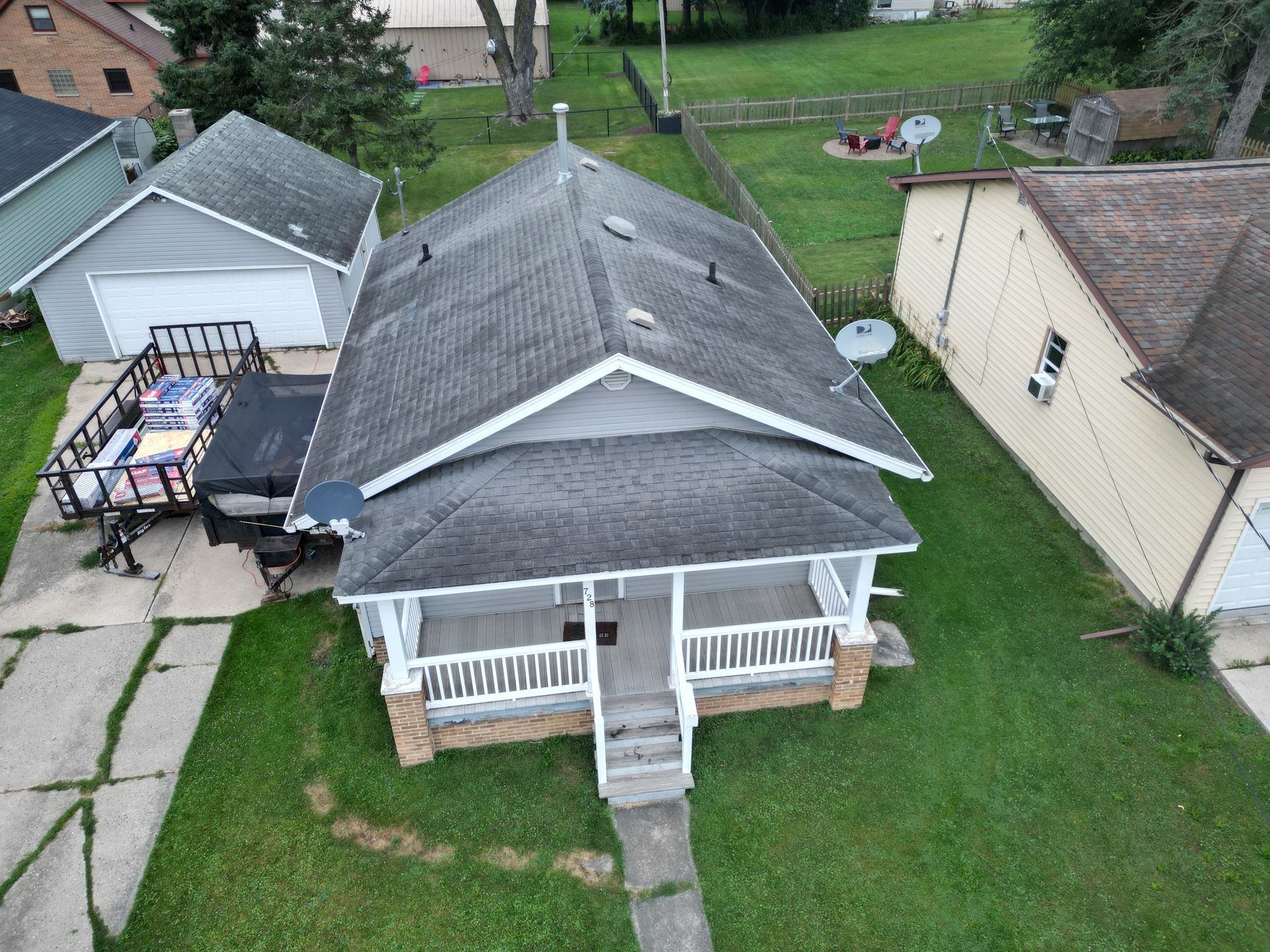 Aerial view of a small gray house with a front porch, grassy yard, and neighboring homes.