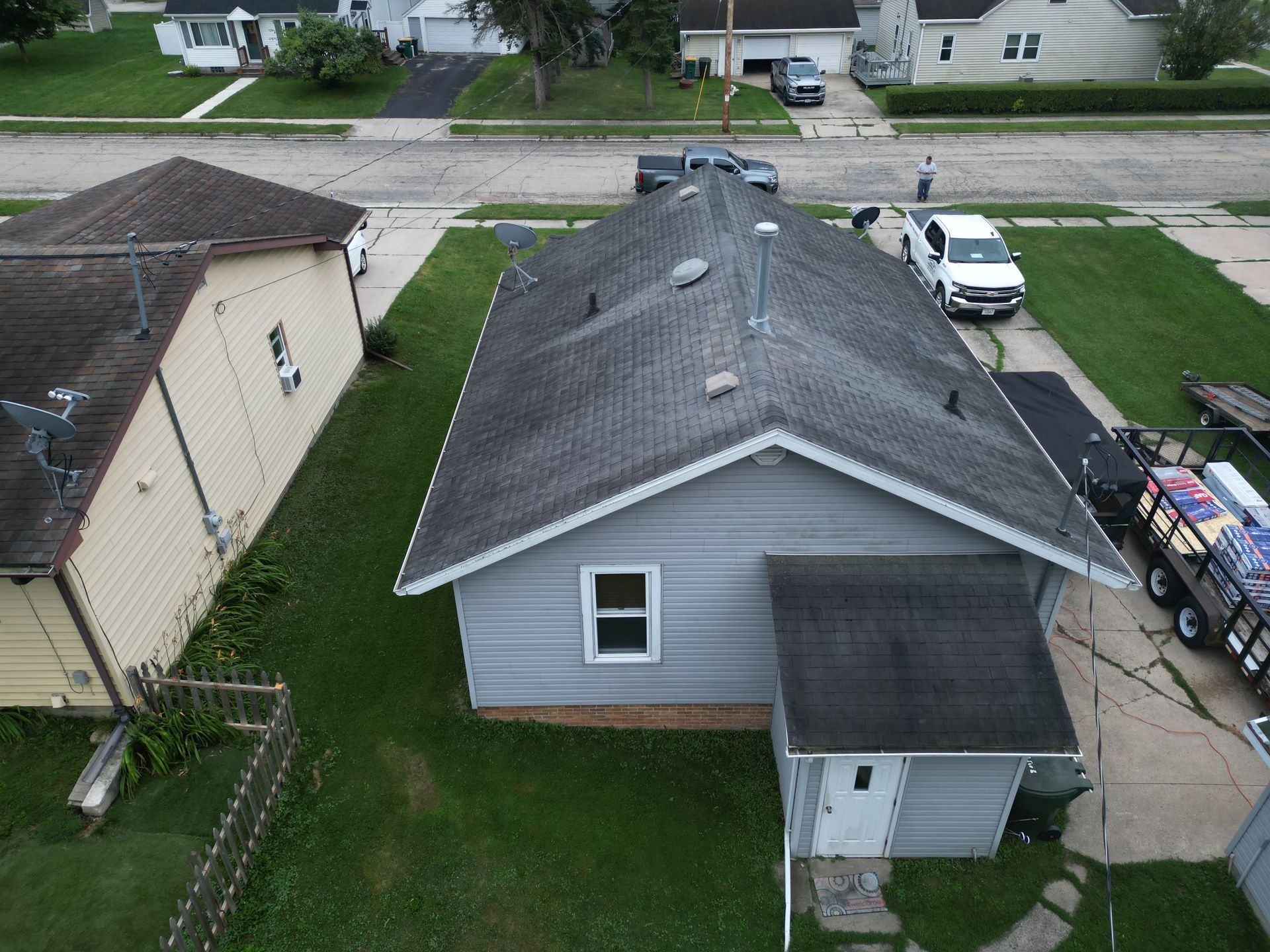 Aerial view of a gray house with a dark roof, driveway, and parked vehicles in a suburban neighborhood.