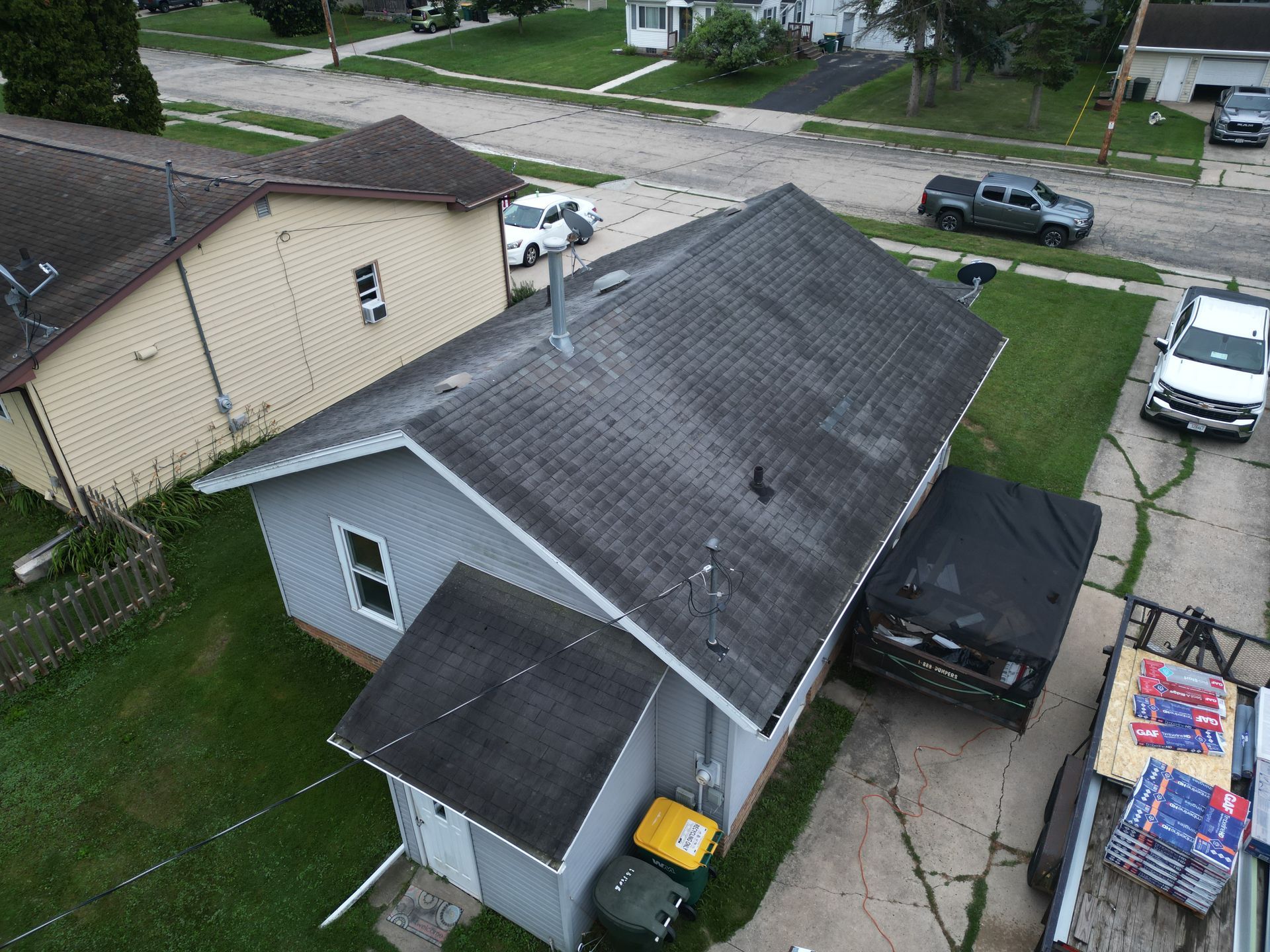 Aerial view of a gray house with a black roof and driveway, beside a parked car and a dumpster trailer.