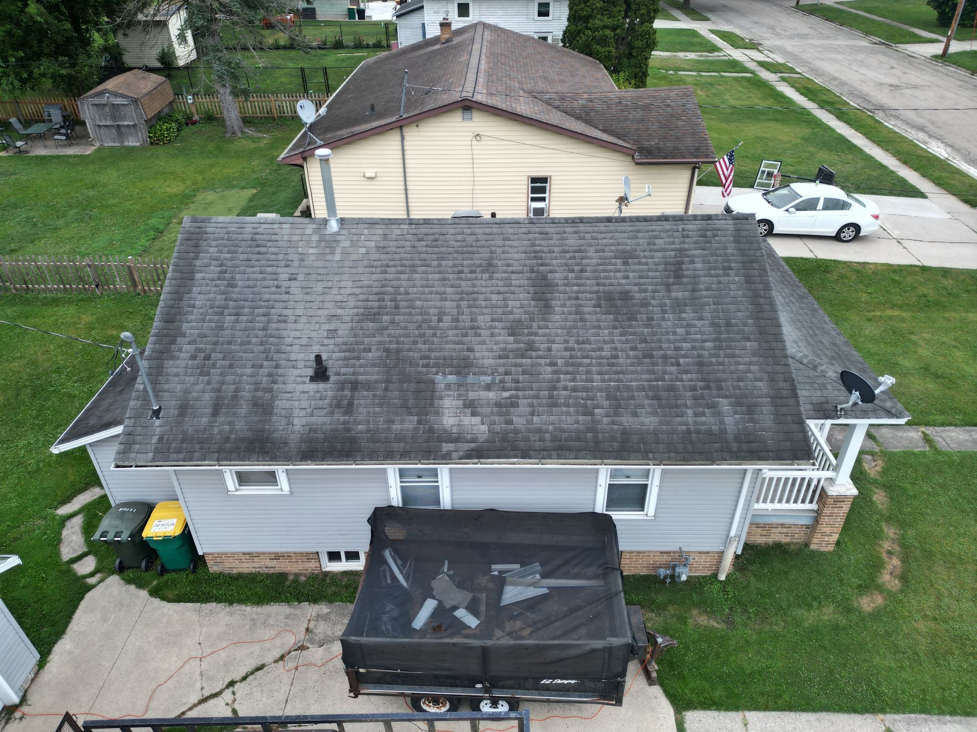 Aerial view of two houses with gray roofs, a black dumpster, and parked vehicles in a residential yard