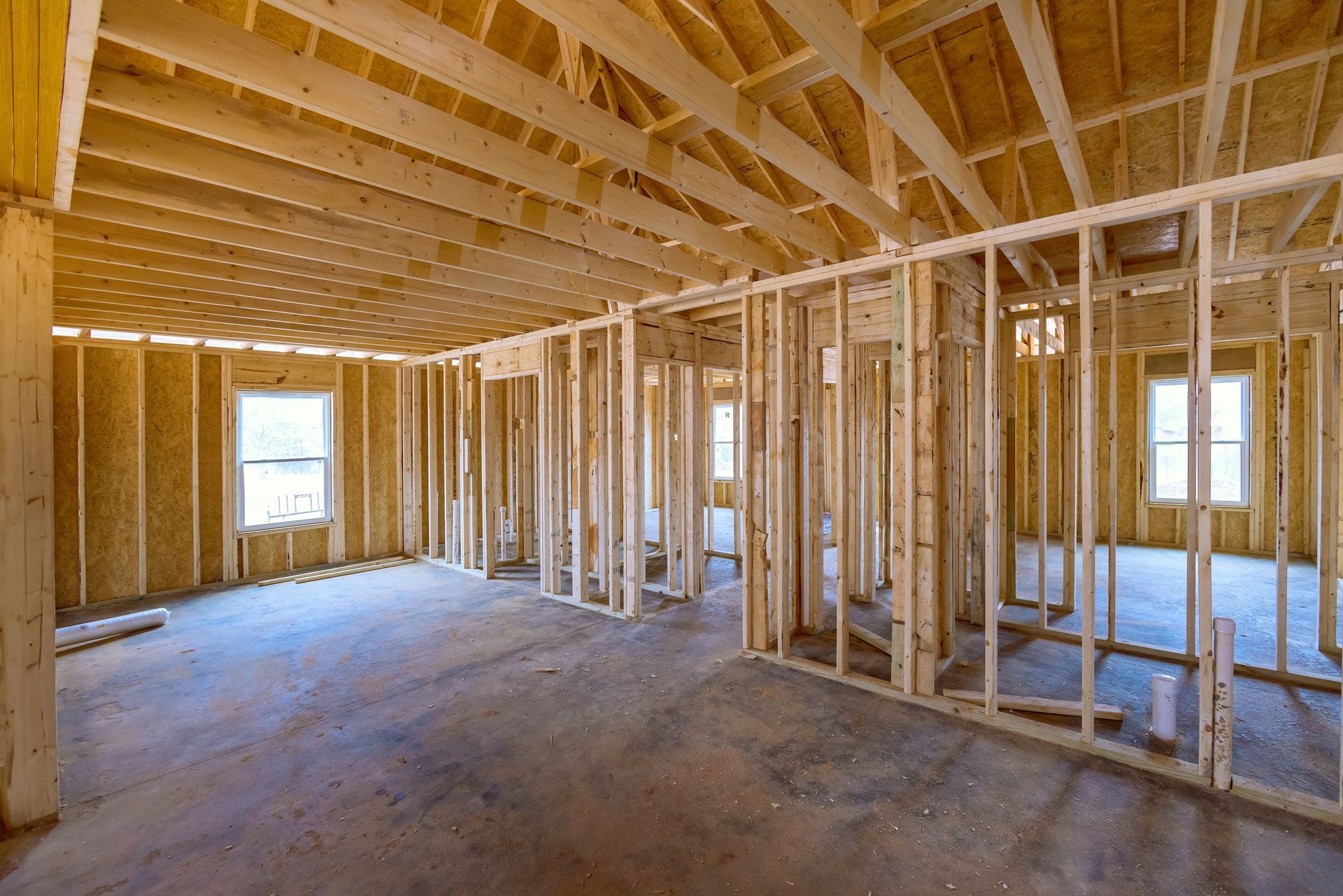 Framed house interior with exposed wooden studs, trusses, and unfinished concrete floor.