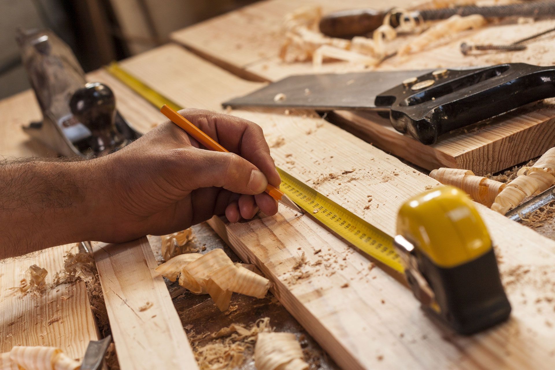 Hand using a pencil and tape measure on a wooden workbench with sawdust and tools