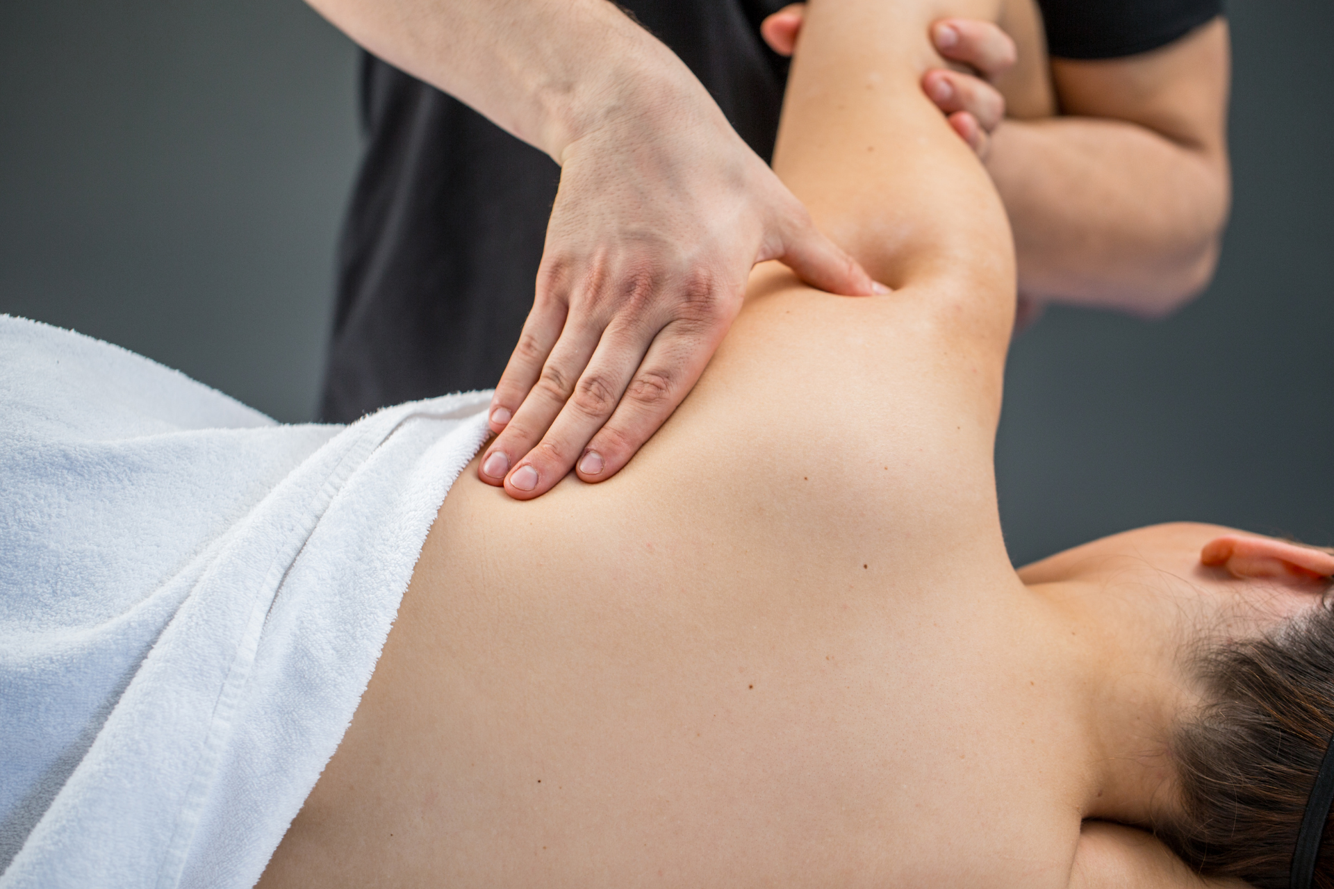Person receiving a shoulder massage from a masseuse, in a room, on a white towel.