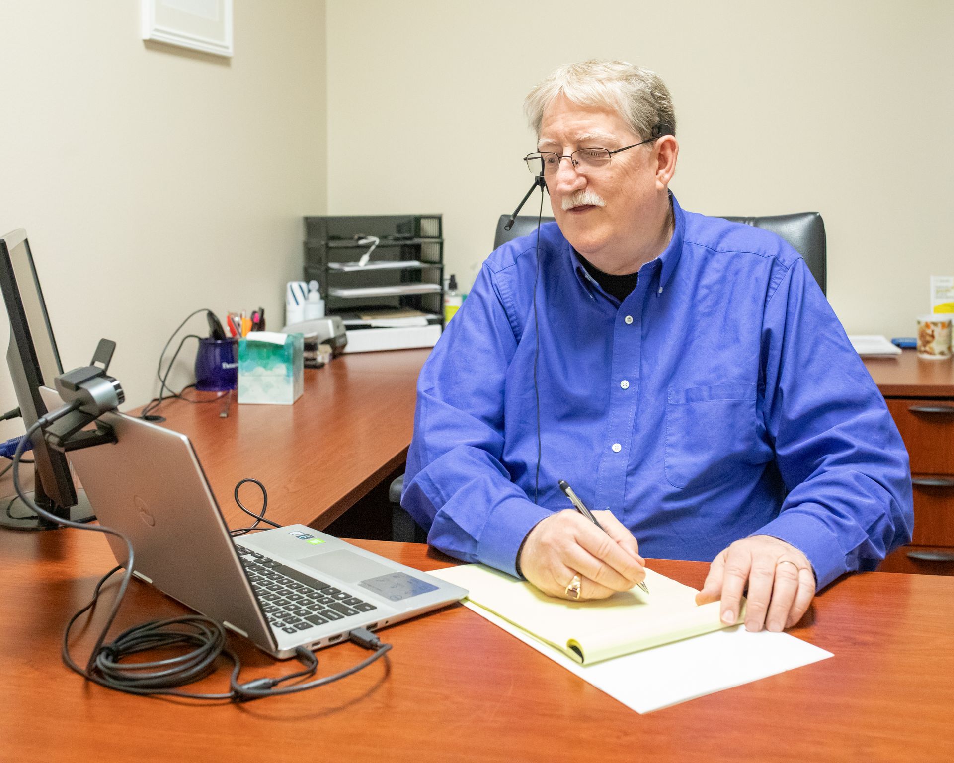 Man at desk taking notes during a video call on a laptop. He's wearing a blue shirt and glasses. Office setting.