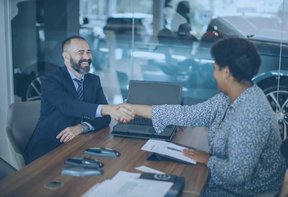 Um homem de terno aperta a mão de uma mulher em uma mesa dentro de uma concessionária de automóveis.