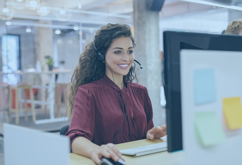 Mulher de cabelos cacheados, usando fones de ouvido, sorrindo e trabalhando em um computador em um escritório moderno.
