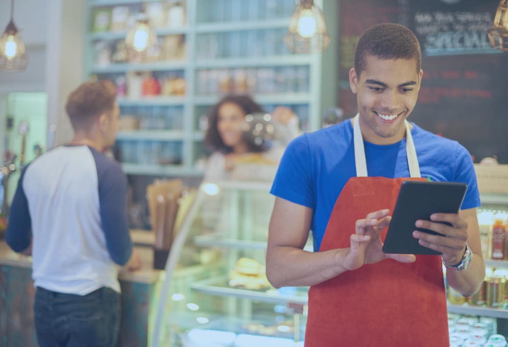 Homem de avental vermelho usando um tablet em um café, sorrindo. Outras pessoas e a vitrine de alimentos estão visíveis.