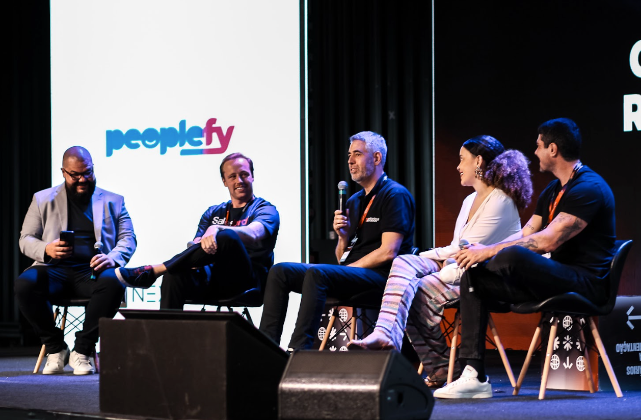 Painel com cinco palestrantes em um palco com o logotipo 