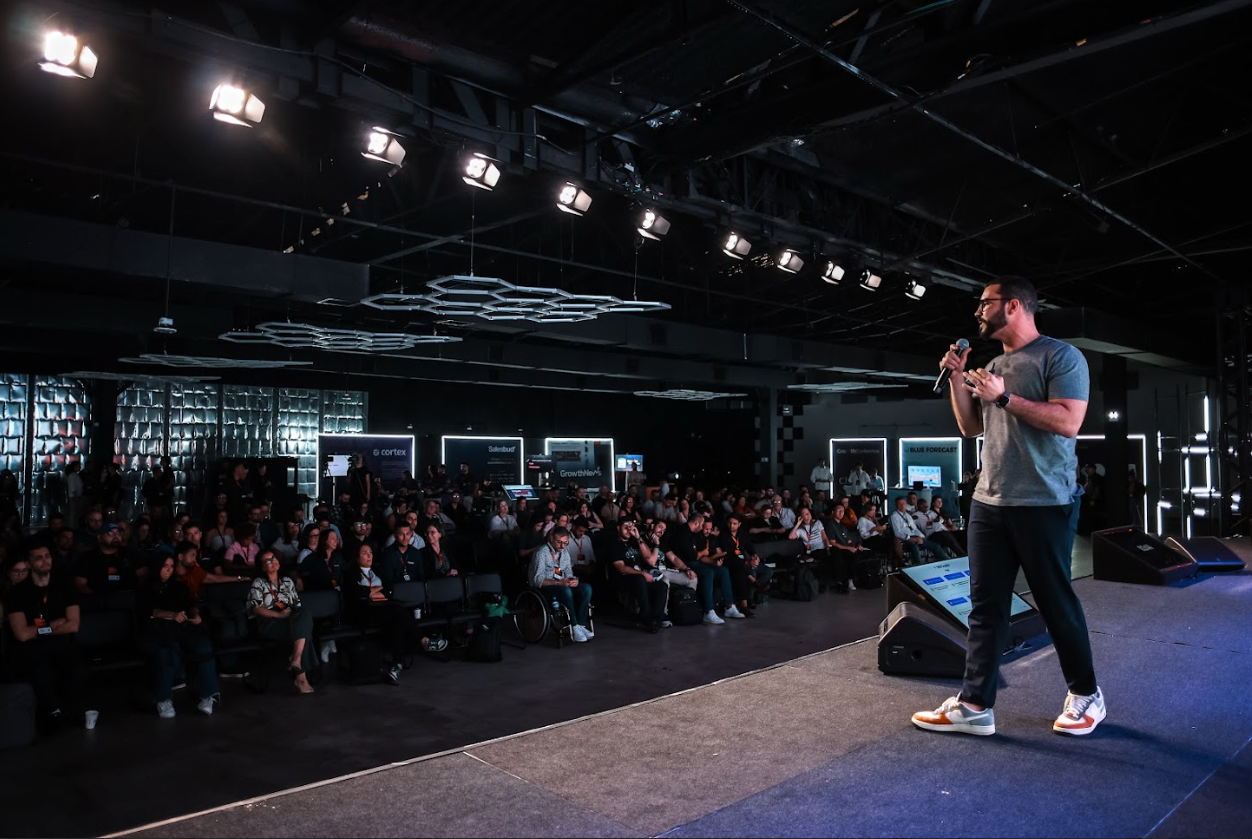Homem falando ao microfone no palco para uma plateia sentada em um evento.