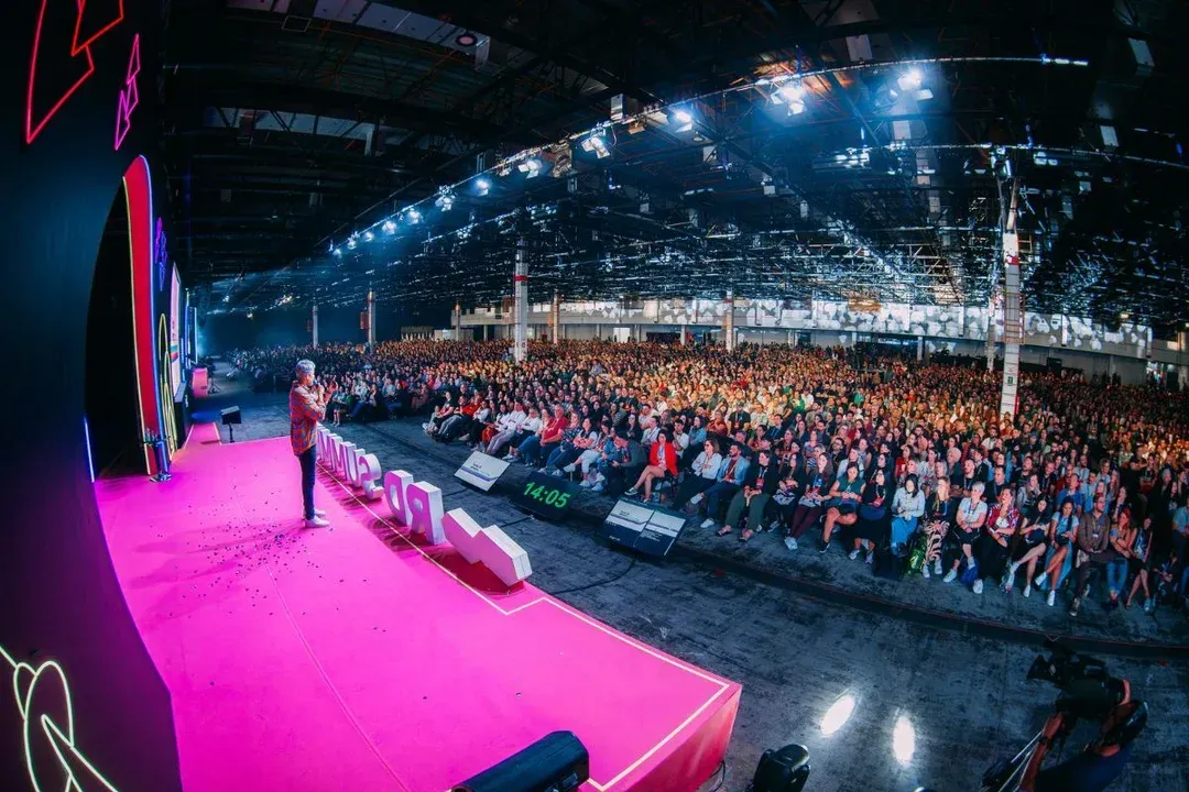 Uma pessoa se dirige a uma grande plateia em um palco bem iluminado. Palco rosa, fundo escuro.