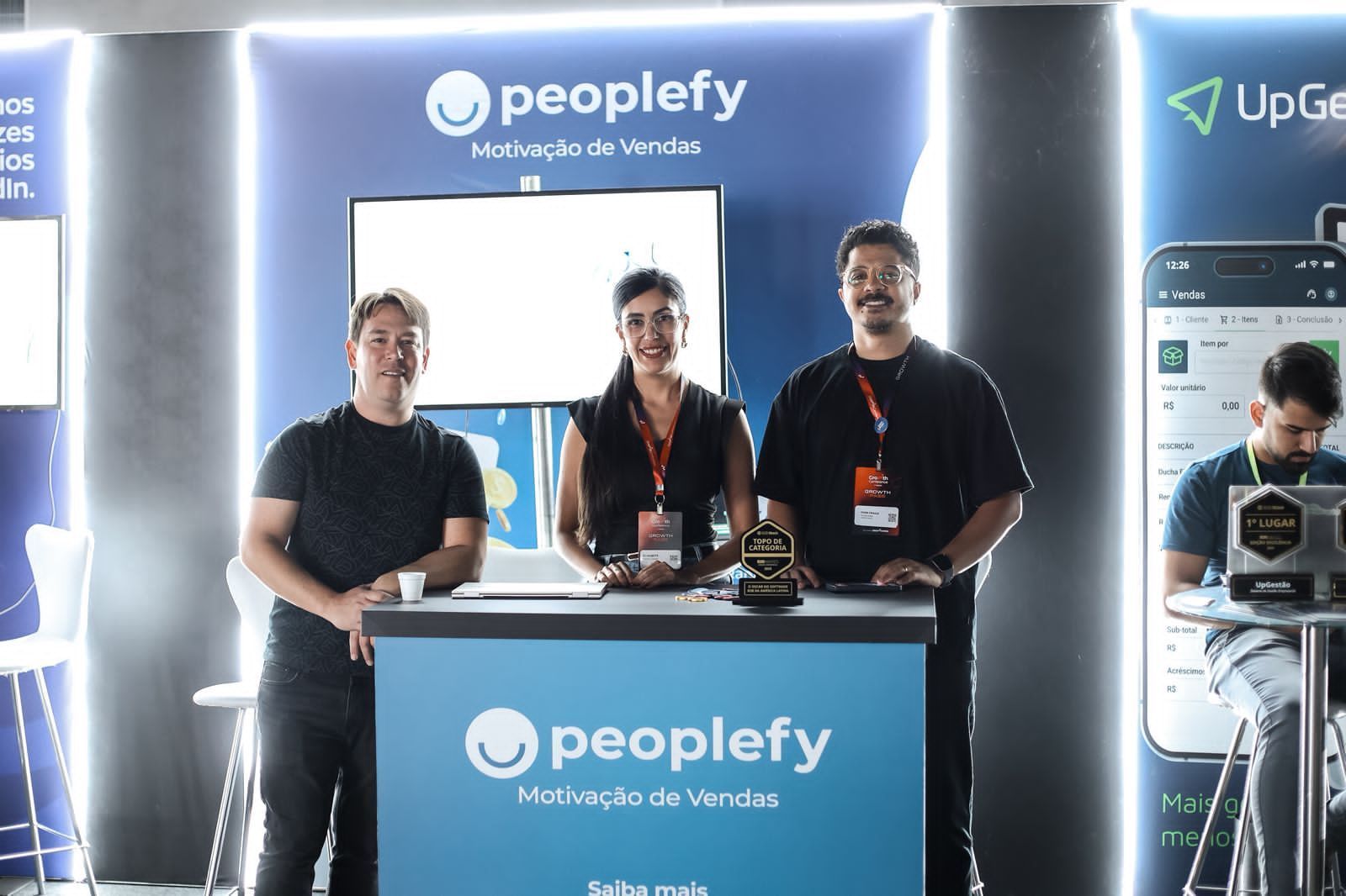 Equipe de vendas da Peoplefy em um estande com uma tela e um logotipo; duas pessoas sorrindo para a câmera.