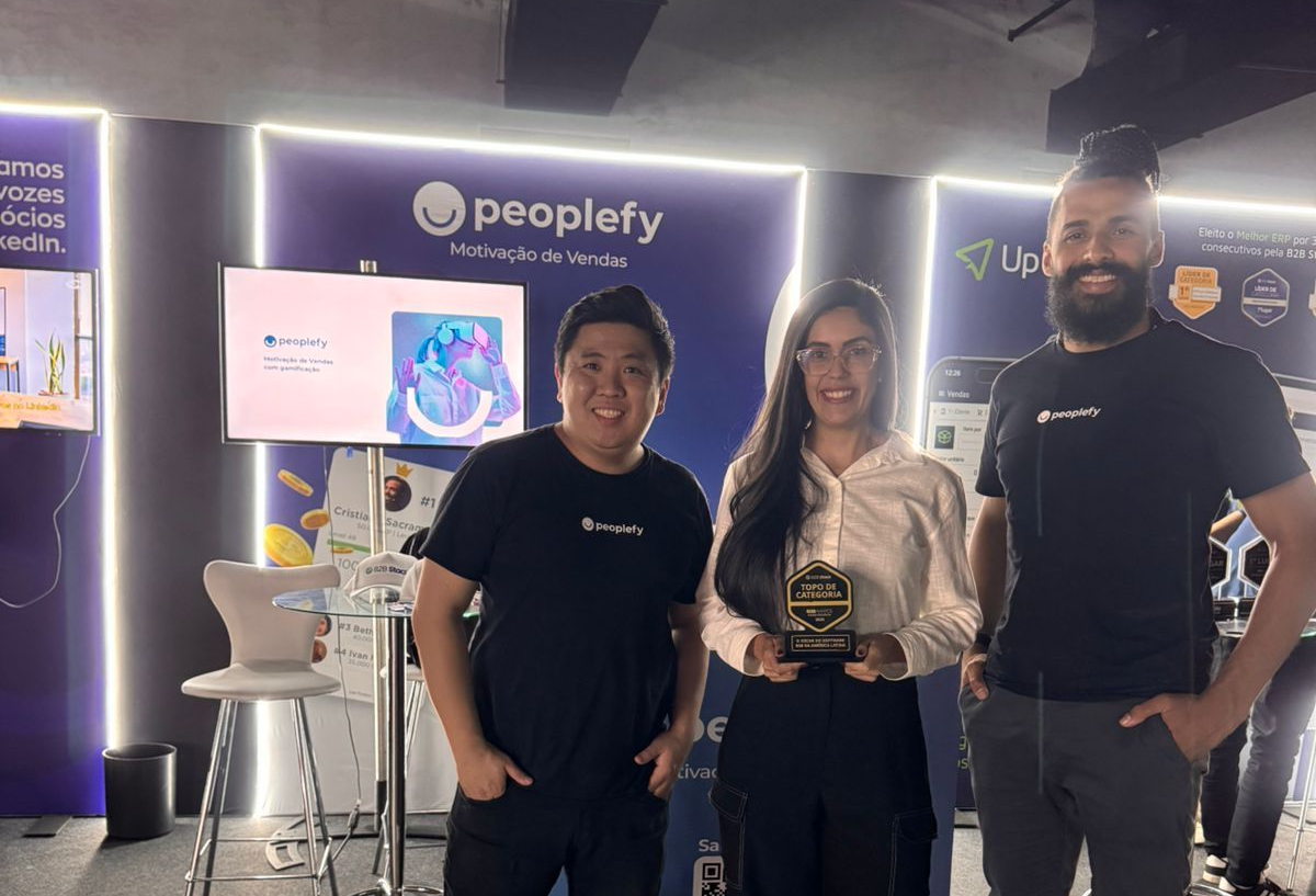 Três pessoas estão sorrindo e segurando um prêmio. Elas estão em frente a um painel de fundo da Peoplefy.
