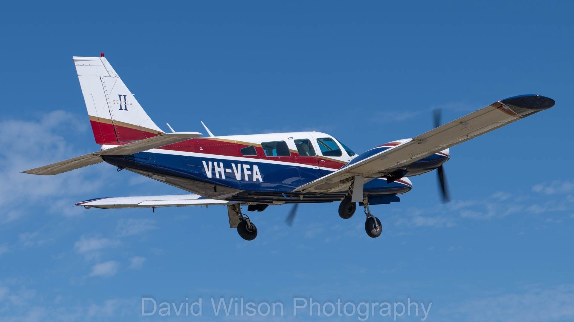 Small airplane, white, blue, and red, in flight against a blue sky; registration VH-VFA visible.
