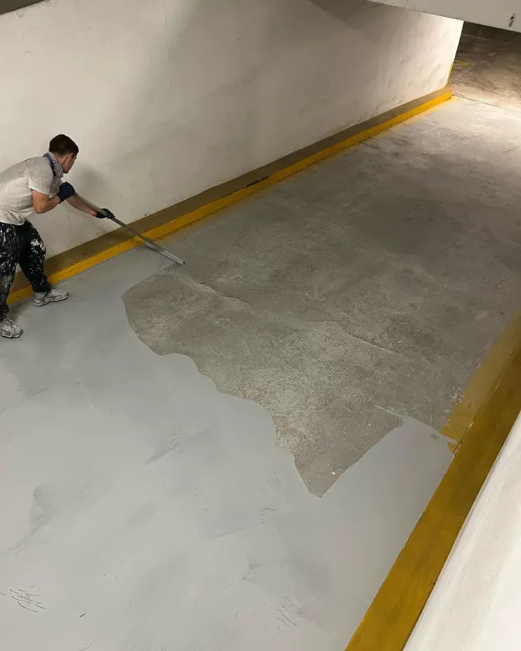 Worker applying white coating to a concrete floor in a parking garage stairwell area