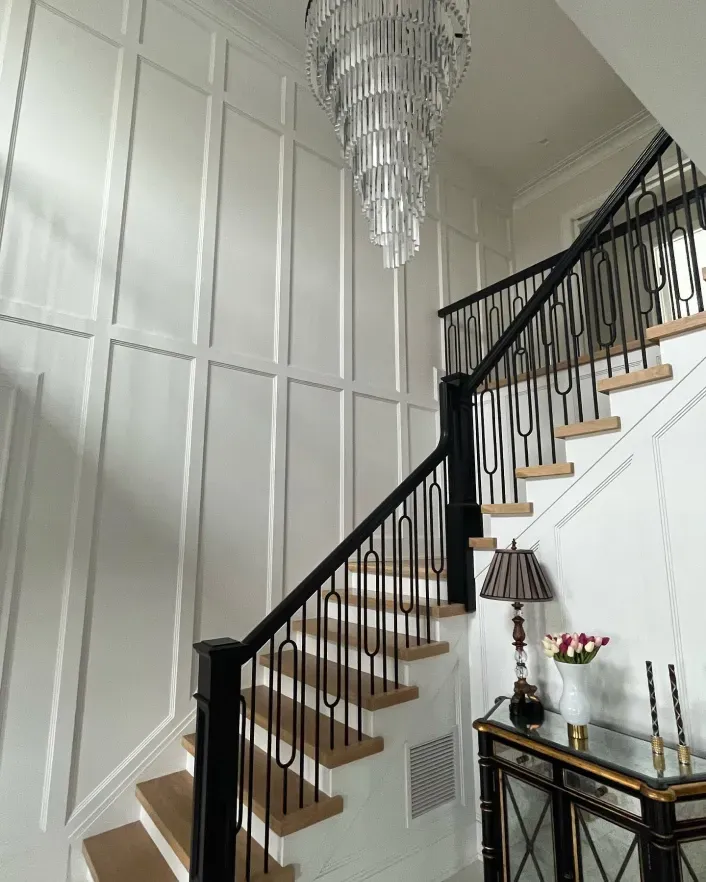 Elegant staircase with black railing, white paneled walls, and a crystal chandelier in a bright entryway
