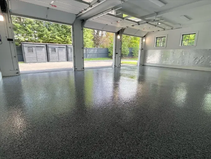 Empty garage with glossy black epoxy floor and open doors to a driveway and greenery.