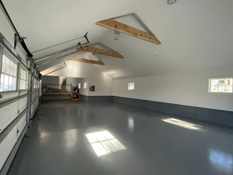 Empty garage interior with white walls, gray floor, and overhead storage rafters
