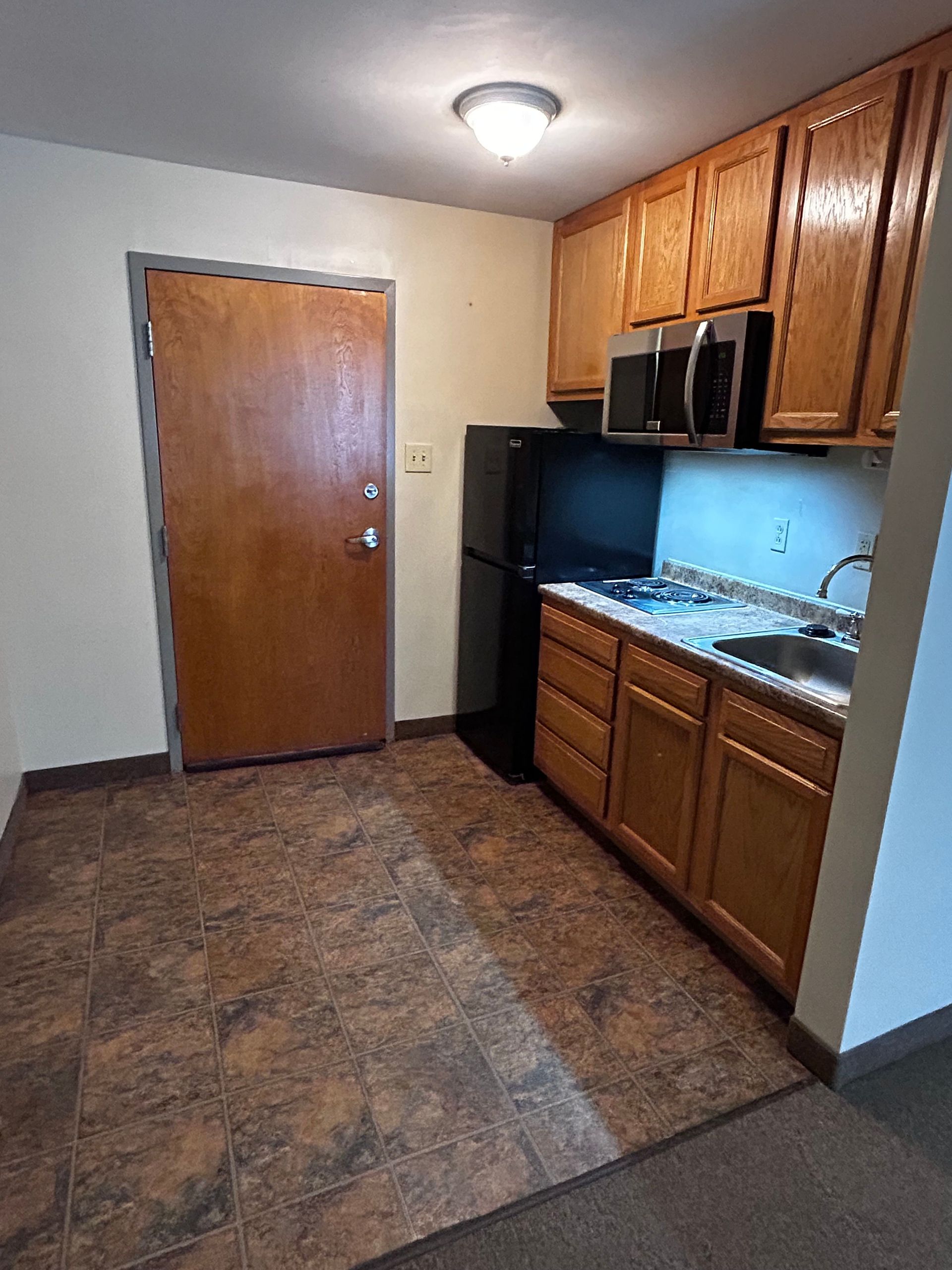 Small kitchen with wood cabinets, black appliances, tiled floor, and a closed door.
