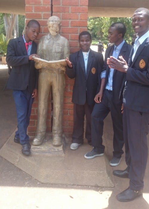 Four Gracious students in dark blazers posing beside a statue in front of a brick wall.