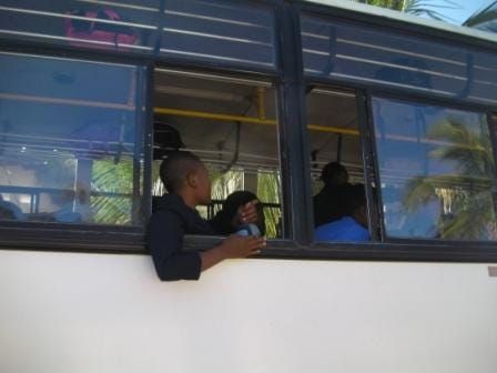 Gracious students on a bus, one by an open window with his arm laying on the sill, looking at the other students.
