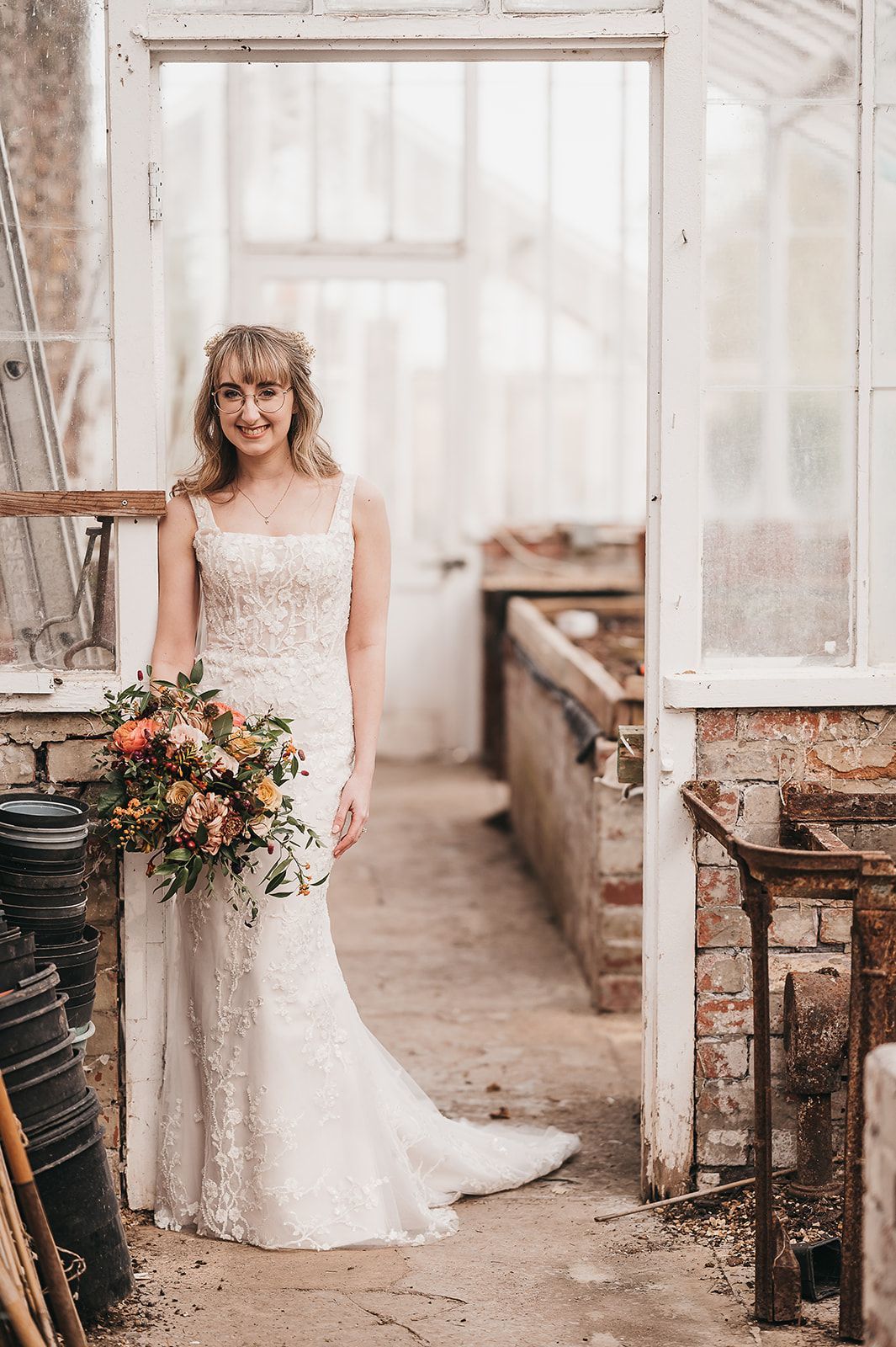 A bride in a wedding dress is standing in a greenhouse holding a bouquet of flowers.
