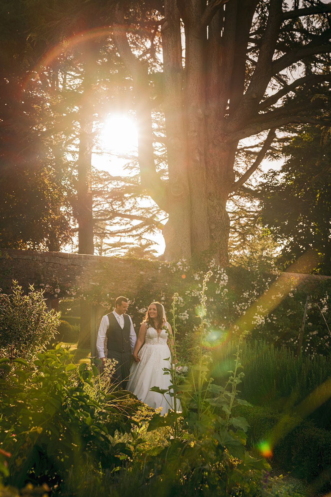A bride and groom are standing in a forest with the sun shining through the trees.