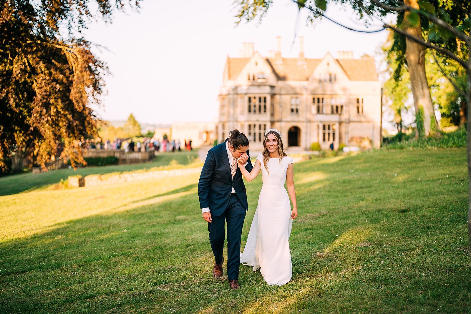 A bride and groom are walking in a field in front of a large house.