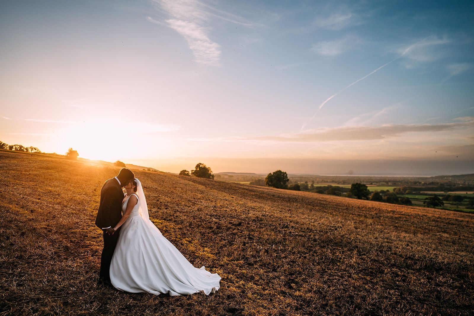A bride and groom are kissing in a field at sunset.