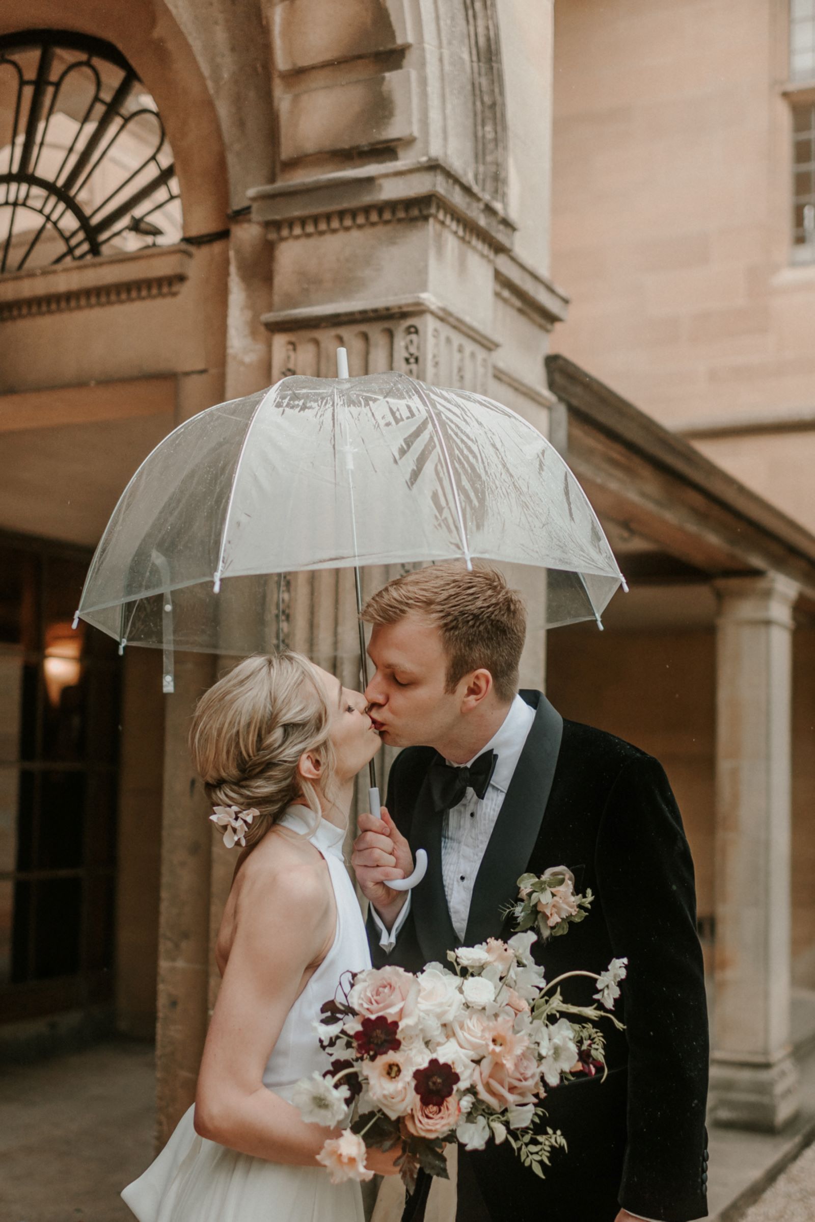 A bride and groom are kissing under an umbrella in the rain.