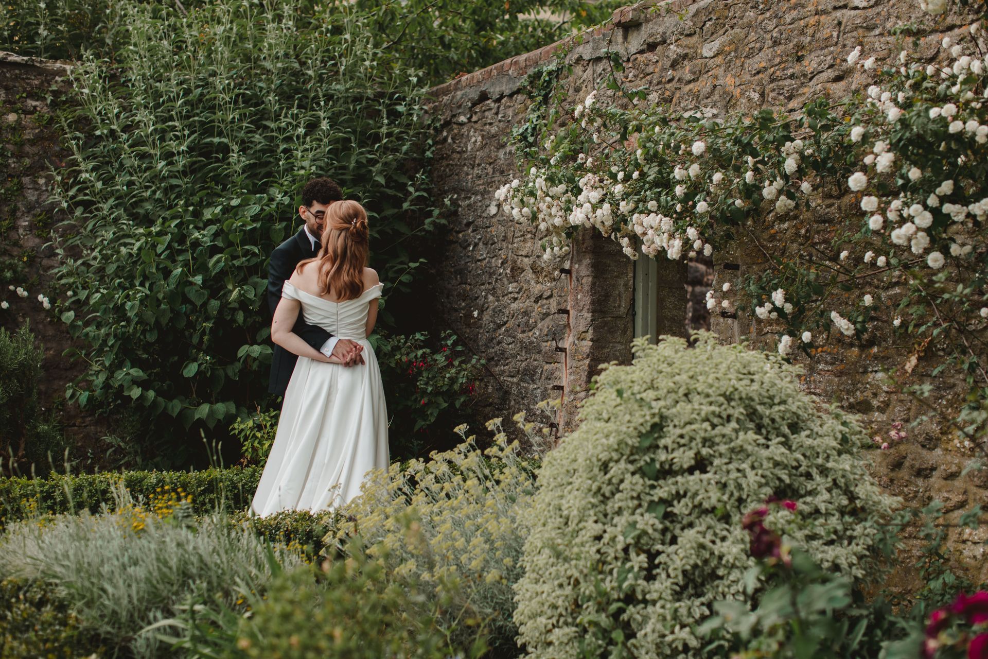 A bride and groom are hugging in a garden in front of a stone wall.
