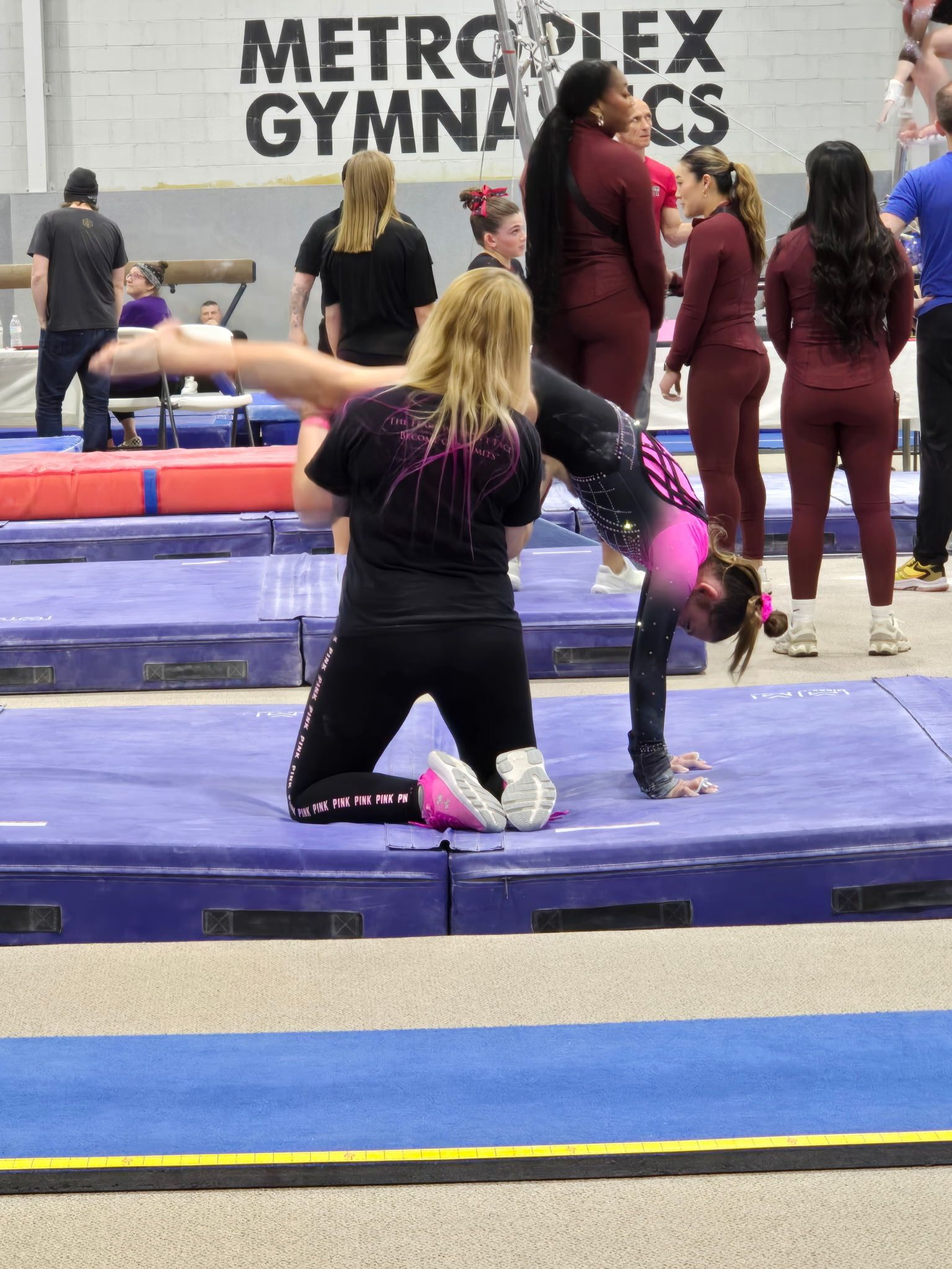 Gymnast in pink leotard doing handstand with coach at competition; indoor gym setting.
