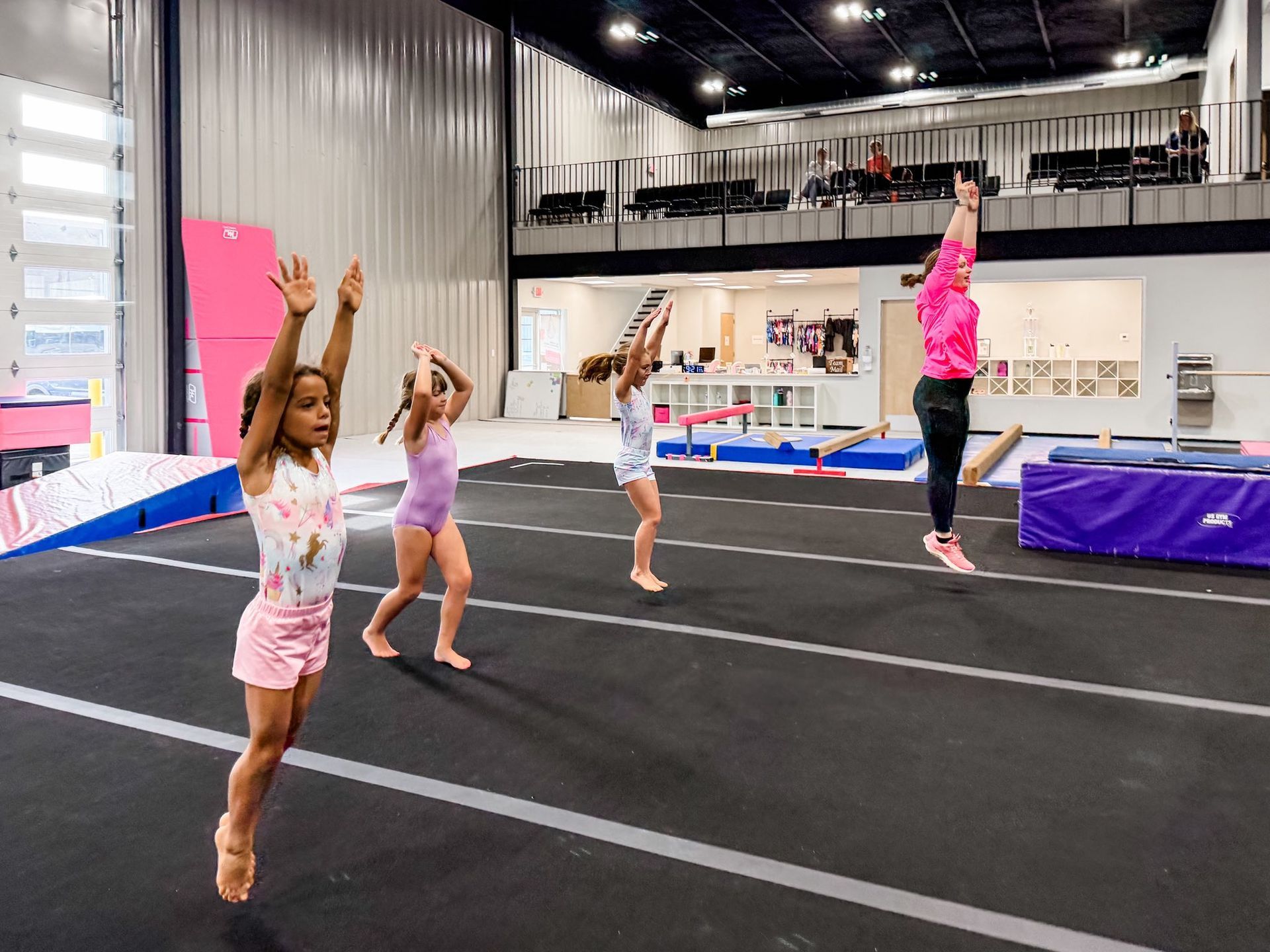 Girls jumping in a gymnastics class; coach watching. Gymnasium with black mat, pink outfits.
